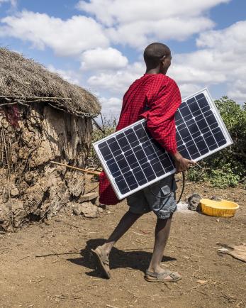 man carrying a solar panel near a hut