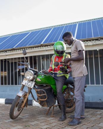 Two men, one standing over a motorcycle, look at a phone together with a solar array behind them.