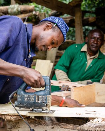 Man using jigsaw to cut plywood, while another man looks on.